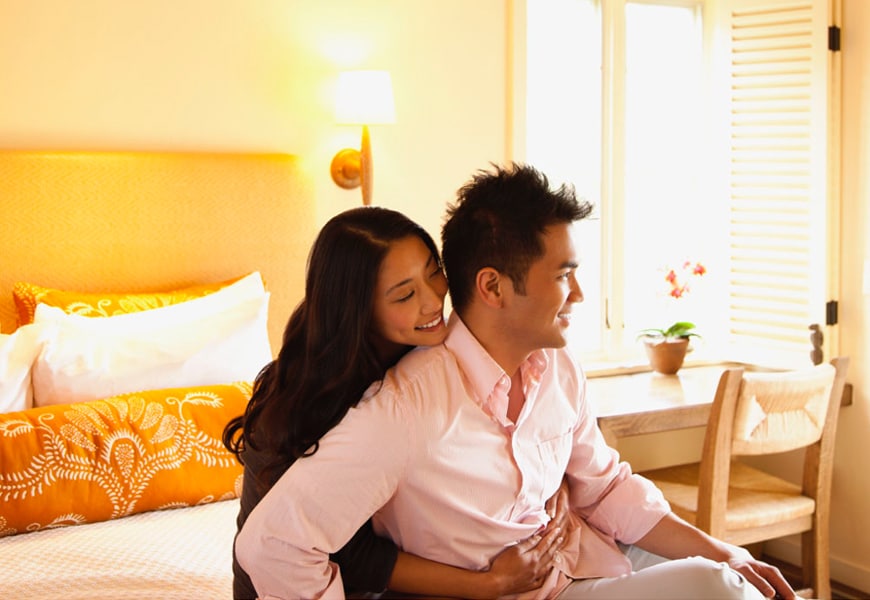 woman wrapping arms around a man both smiling on the bed with lights on and windows open